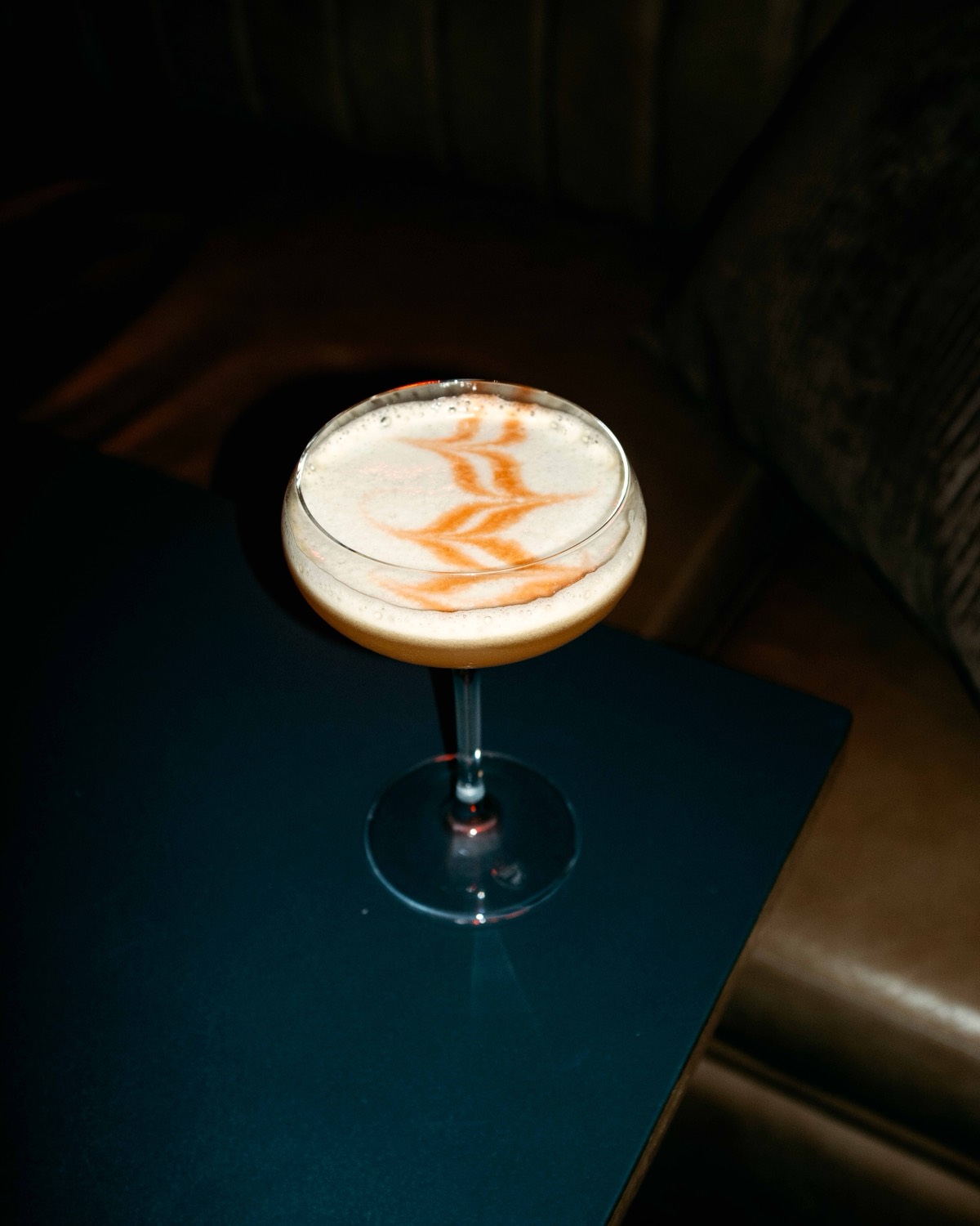 En cocktail i coupeglas med mönstrat skum, på ett mörkt sidobord framför en grön sammetssoffa.