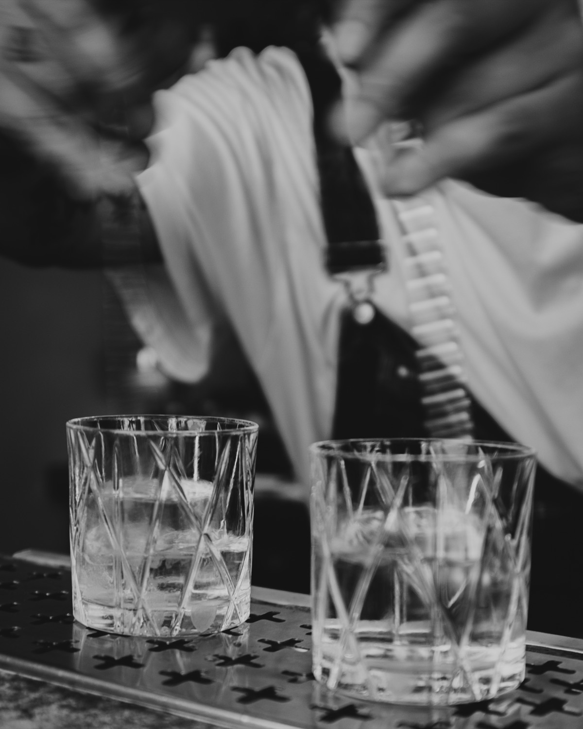 En bartender silar upp en drink i två slipade glas, i svartvitt.